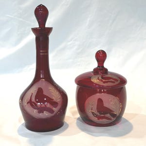 May include: A ruby red glass decanter with a matching stopper and a lidded glass jar. Both pieces feature etched circular panels displaying a bird perched on a branch, accented with gold detailing. The glass has a deep, translucent crimson hue.
