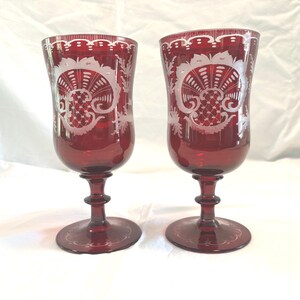 May include: A pair of deep red glass goblets featuring intricate white etched patterns around the upper bowls. Each stemware piece has a clear, circular base and a slender stem leading to a rounded bowl with detailed floral and lattice engravings.