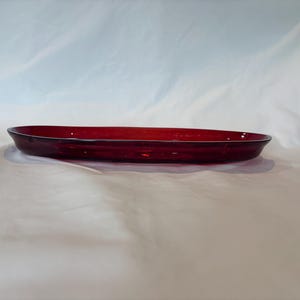 May include: A translucent, deep red glass tray with an elongated oval shape. The tray features smooth, curved edges and a shallow basin, showcasing a vibrant crimson hue that catches the light across its polished, reflective surface.