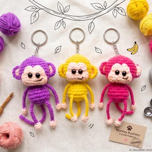 May include: Three crocheted monkey keychains in purple, yellow, and pink, each with a metal ring, black plastic eyes, and light beige faces. The set includes yarn balls, a wooden crochet hook, and a card labelled Monkey Business Crochet Patterns.