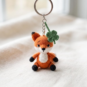 Op de afbeelding: Een gehaakte vossen-sleutelhanger met een kleine oranje en witte vos met zwarte pootjes en oren, bevestigd aan een zilveren metalen ring. Een klein gehaakt klavertjevier is bevestigd aan de ketting boven het figuurtje van de vos.