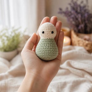 May include: A small, round, handmade crochet doll featuring a cream-coloured head and a sage green body. The doll has two small, black, circular bead eyes positioned on the upper cream section. The texture is consistent with yarn stitching.