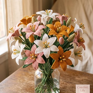 May include: A bouquet of crocheted lily flowers in white, soft pink, and golden yellow, featuring detailed petals, yellow stamens, and green stems, arranged in a clear glass vase. A small square label in the corner displays the text Comfort Lily.