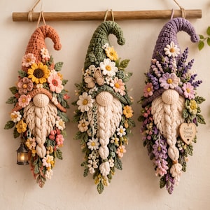 May include: Three crocheted gnome wall hangings suspended from a wooden rod. The left gnome is orange with sunflowers, the center is green with daisies and a butterfly, and the right is purple with lavender and a heart tag reading Good Things Grow.