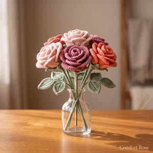 May include: A bouquet of crocheted roses in shades of pink, mauve, and coral, featuring detailed petals and green crocheted leaves on stems. The flowers are arranged in a clear glass vase. The text Comfort Rose is visible in the bottom right corner.