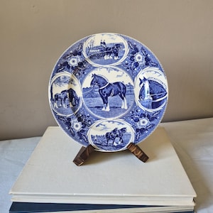 May include: A round ceramic plate featuring a blue and white transferware pattern. The design includes a central circular panel depicting a draft horse, surrounded by four smaller circular vignettes showing horses in various scenes, all set against a floral background.