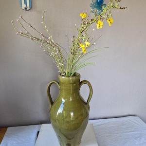 May include: A glossy olive green ceramic vase with two side handles, containing a bouquet of thin green stems with small pink buds and bright yellow flowers. The vase is positioned on top of a stack of two books with white and dark blue covers.
