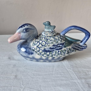 Op de afbeelding: Een keramische theepot in de vorm van een eend met een handgeschilderd blauw-wit schubbenpatroon op het lichaam. De eend heeft een lichtroze snavel, een blauw handvat aan de achterkant en een kleine eendvormige knop op het afneembare deksel.