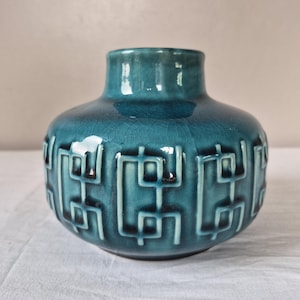 May include: A teal ceramic vase with a glossy crackle glaze finish. The body of the vase features a repeating embossed geometric pattern of interconnected rectangular lines encircling the center, with a short, wide neck and a rounded base.
