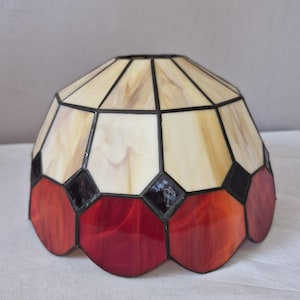 Könnte beinhalten: Ein kuppelförmiger Lampenschirm aus Buntglas mit einem geometrischen Muster aus cremefarbenen Paneelen im oberen Bereich und einer Reihe tiefroter, bogenförmiger Paneele an der Basis, die durch dunkle Metalllötstellen verbunden sind.