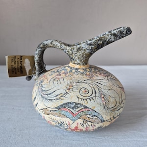 May include: A ceramic pitcher with a rounded body and a single handle, featuring a beige base with intricate black, red, and blue painted patterns. A rectangular tag is attached to the handle with the text: AΦΟΙ ΒΑΡΔΑΚΑΚΗ Α. ΙΩΝΙΑΣ 204 ΗΡΑΚΛΕΙΟ ΚΡΗΤΗΣ ΤΗΛ. 237482.