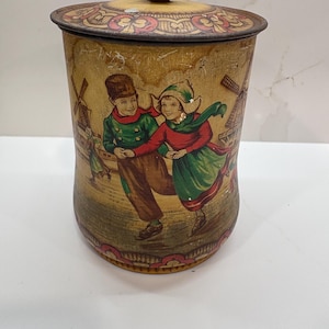 May include: A cylindrical metal canister with a removable lid and a dark brown knob. The exterior features a vintage-style illustration of two children ice skating in a landscape with windmills, rendered in muted yellow, green, and red tones.