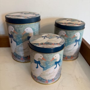 May include: A set of three cylindrical metal canisters of varying heights, each featuring a light blue background with white geese wearing blue bows. The lids are dark blue, and the geese are depicted in a snowy setting on the sides and tops.
