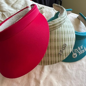 May include: Three sun visors arranged in a row. The first is solid bright pink. The second is a light green and white plaid pattern with the text IZOD embroidered on the front. The third is solid teal with the TaylorMade logo and text.