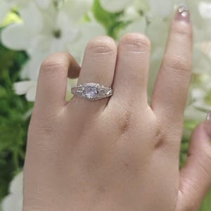 May include: A silver-toned ring featuring a central round clear stone flanked by smaller accent stones on each side. The band displays intricate metalwork detailing along the shank, creating a textured appearance around the finger.
