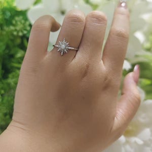 May include: A silver-toned ring featuring a central round white opal stone surrounded by a sunburst design of small clear stones. The band is thin and metallic, with the starburst setting extending outward from the center stone in a symmetrical pattern.