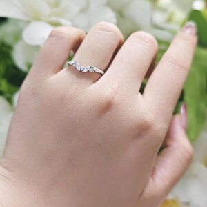 May include: A silver-toned chevron ring featuring a series of small, clear, sparkling stones set along the V-shaped band. The ring has a polished metallic finish and follows a curved, pointed design that sits across the finger.