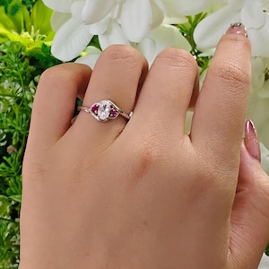 May include: A silver-toned ring featuring a central oval-shaped clear stone flanked by two smaller round pink stones. The band has a bypass design that curves around the stones, creating a delicate and polished appearance on the finger.