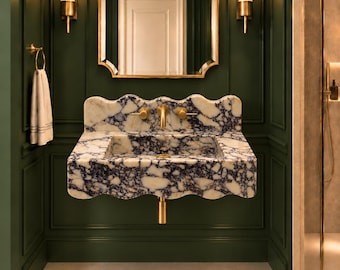 May include: A wall-mounted bathroom sink crafted from white and dark purple marble with a scalloped edge design. The sink features gold-toned metal faucets and a matching drain pipe, set against dark green panelled walls with gold wall sconces.