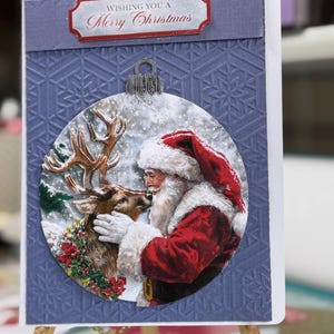 May include: A rectangular greeting card with a textured periwinkle blue background featuring a debossed snowflake pattern. A circular ornament cutout displays an illustration of Santa Claus with a reindeer. The text reads, Wishing You A Merry Christmas.