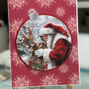 May include: A rectangular greeting card with a deep red background featuring a white snowflake pattern. A circular cutout displays an illustration of Santa Claus holding a reindeer, topped with a silver ornament cap to resemble a Christmas bauble.