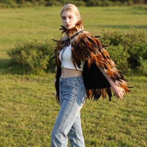May include: A cropped white sleeveless top, light blue denim jeans, and a layered feather cape featuring long, pointed feathers in shades of brown, black, and tan. The cape is secured at the neck with a thin black ribbon tie and has a black lining.