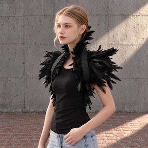 May include: A black feather shrug featuring a high, structured collar and layered plumage that extends over the shoulders. The garment is worn over a simple black tank top and light blue denim jeans, showcasing a textured, dark aesthetic.