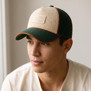 Puede incluir: Una gorra de béisbol con un panel frontal color crema que incluye un parche rectangular, una visera verde oscuro con borde naranja y un panel trasero de malla verde oscuro. La gorra presenta una textura tejida en la parte delantera.