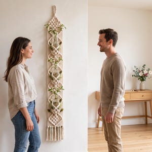 Op de afbeelding: Een lange, verticale macramé wandhanger van off-white koord, voorzien van drie geïntegreerde glazen reageerbuishouders. Elke glazen buis bevat een kleine groene plantstek. Het macraméontwerp bevat ingewikkelde knopen en een onderkant met franjes.