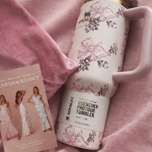 Puede incluir: Un vaso Stanley Quencher ProTour de color rosa p&aacute;lido con capacidad de 1,18 L, con un estampado de flores y lazos, borde dorado y asa rosa a juego. Una tarjeta rectangular con el texto LoveShackFancy e im&aacute;genes de tres vestidos.