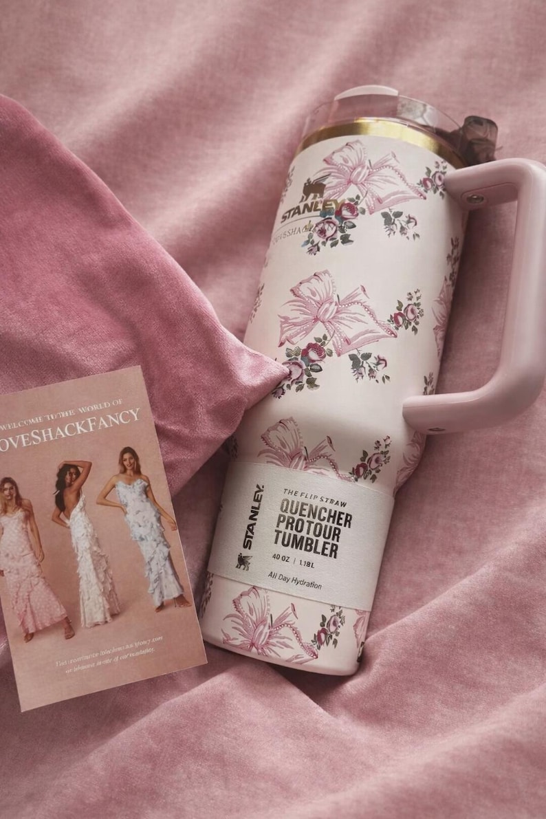 Puede incluir: Un vaso Stanley Quencher ProTour de color rosa p&aacute;lido con capacidad de 1,18 L, con un estampado de flores y lazos, borde dorado y asa rosa a juego. Una tarjeta rectangular con el texto LoveShackFancy e im&aacute;genes de tres vestidos.