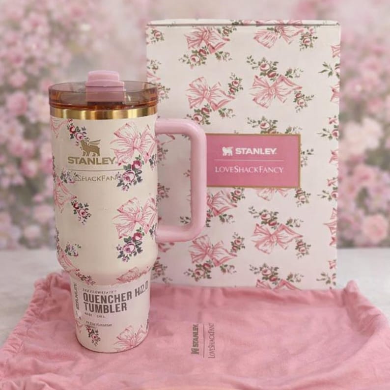 Puede incluir: Un vaso Stanley Quencher H2.0 de color crema con un estampado de flores y lazos rosas, un asa rosa a juego y una tapa transparente con un tap&oacute;n rosa. El vaso se muestra junto a su caja a juego y una bolsa de tela rosa con cord&oacute;n.