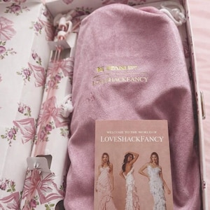 Puede incluir: Una caja de regalo rectangular con estampado floral que contiene una bolsa de tela rosa con el texto dorado STANLEY LOVESHACKFANCY. Dentro hay una pajita rosa larga y una tarjeta titulada WELCOME TO THE WORLD OF LOVESHACKFANCY con tres modelos en vestidos.