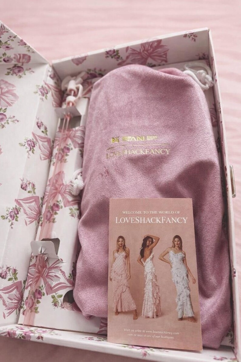 Puede incluir: Una caja de regalo rectangular con estampado floral que contiene una bolsa de tela rosa con el texto dorado STANLEY LOVESHACKFANCY. Dentro hay una pajita rosa larga y una tarjeta titulada WELCOME TO THE WORLD OF LOVESHACKFANCY con tres modelos en vestidos.