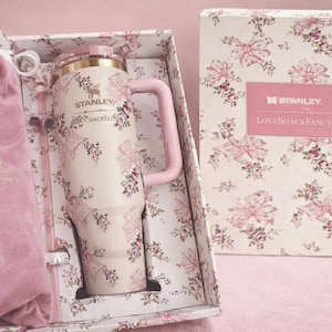 Puede incluir: Un vaso Stanley de color crema con asa rosa y un patr&oacute;n de flores y lazos, que presenta el texto Stanley y LoveShackFancy. Se presenta en una caja floral a juego con una bolsa de terciopelo rosa que tambi&eacute;n lleva los nombres de la marca.