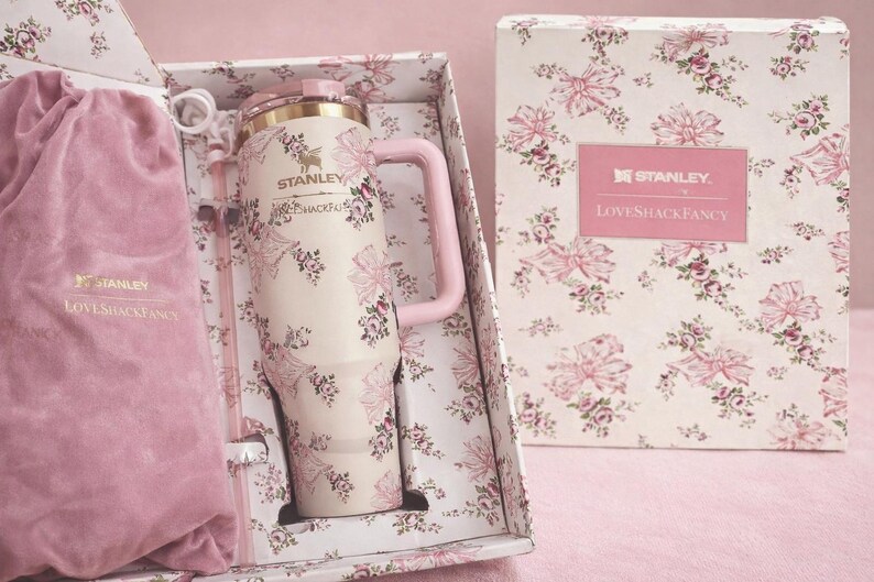 Puede incluir: Un vaso Stanley de color crema con asa rosa y un patr&oacute;n de flores y lazos, que presenta el texto Stanley y LoveShackFancy. Se presenta en una caja floral a juego con una bolsa de terciopelo rosa que tambi&eacute;n lleva los nombres de la marca.