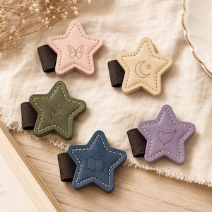 May include: Five star-shaped pen holders with elastic bands. Each features a unique embossed icon: a butterfly on pink, a crescent moon on beige, a leafy branch on olive green, an open book on navy blue, and a heart on light purple leather.