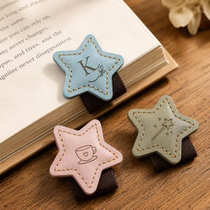 May include: Three star-shaped bookmarks with brown elastic bands. One light blue star features the letter K and a small flower, one light pink star shows a teacup with a heart, and one sage green star displays a magic wand with sparkles.
