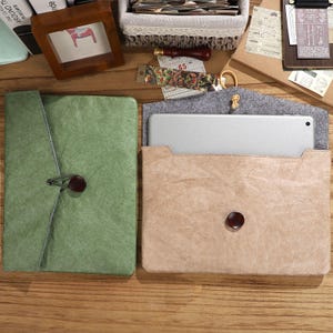 May include: Two rectangular protective sleeves for electronic tablets. One sleeve is sage green with a dark brown button closure and elastic cord. The other sleeve is tan with a dark brown button closure, containing a silver tablet inside.