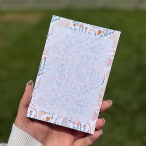 Pode incluir: Um bloco de notas retangular com um padrão repetitivo de gansos brancos, folhagem azul-clara e pequenas flores cor de laranja sobre um fundo branco. O design cobre toda a superfície do papel, exibindo uma paleta de cores suaves e delicadas.