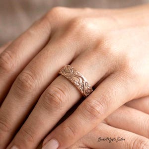 May include: A rose gold band ring featuring an intricate, embossed floral and vine pattern that wraps around the entire circumference of the metal. The surface of the ring has a textured, matte finish with detailed scrollwork and small flower motifs.