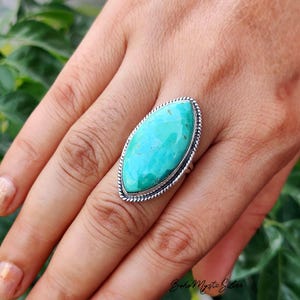 May include: A marquise-shaped turquoise gemstone ring set in a silver band with a twisted rope-style border. The stone is a vibrant teal-green color with natural dark matrix inclusions, securely held in a polished silver setting on a finger.