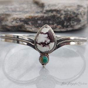 May include: A silver cuff bracelet featuring a central teardrop-shaped white stone with dark brown matrix patterns, accented by a small round turquoise stone below. The band has a twisted rope detail along the centre and a polished finish.