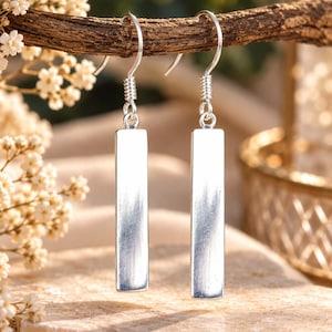 Peut inclure: Une paire de boucles d'oreilles pendantes argentées composées d'un pendentif en forme de barre rectangulaire lisse suspendu à un crochet. La surface métallique présente une finition polie qui reflète la lumière, avec le texte "Boho Mystic Silver" inscrit en bas.