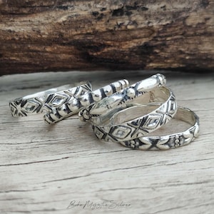 May include: A set of four silver rings featuring various embossed patterns, including geometric diamond shapes, beaded textures, and smooth polished sections. The rings are stacked together, displaying a metallic finish with darkened recessed details.