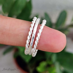 Peut inclure: Un ensemble de trois bagues en argent composé de deux anneaux extérieurs à texture torsadée et d'un anneau central à la finition martelée lisse. Les bagues sont empilées, présentant une surface métallique polie et un ton argenté uniforme.