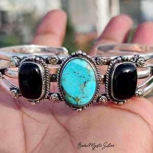 May include: A silver cuff bracelet featuring a central oval turquoise stone flanked by two oval black onyx stones. The metal band has a multi-strand design with twisted wire detailing and small decorative silver beads surrounding each gemstone.