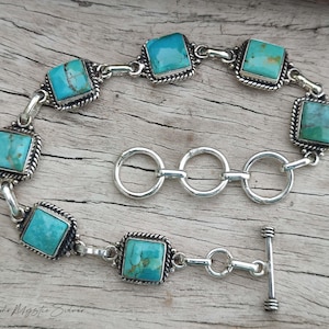 May include: A silver bracelet featuring six square turquoise stones set in twisted metal frames. The bracelet includes a toggle clasp and three circular adjustment rings. The text "Boho Mystic Silver" is printed in the bottom left corner.