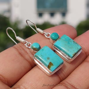 May include: A pair of silver drop earrings featuring a rectangular turquoise stone set above a smaller round turquoise cabochon. The stones display natural brown matrix patterns and are encased in polished silver settings with hook-style wires.