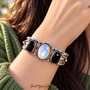 Könnte beinhalten: Ein silberner Armreif mit einem zentralen ovalen Mondstein-Cabochon, flankiert von zwei kleineren ovalen schwarzen Onyxsteinen. Das Metallband weist eine gedrehte Kordelstruktur und dekorative Perlenverzierungen um jeden Edelstein auf.